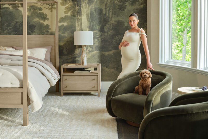 Interiors Photographer Alpha Smoot captures expecting lifestyle expert Olivia Culpo amidst the earthy, lake view inspired decor of her recently redesigned lakeview home bedroom with help from the Crate and Barrel Design Desk. Olivia stands near a window next to a moss green Crate and Barrel velvet chair that seats her small dog.