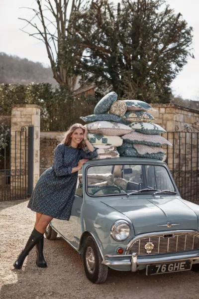 Outdoor lifestyle fashion image with woman in dress and boots leaning on tiny classic car with matching decorative pillows piled on top for Garnet Hill with prop styling by Elkie Brown, represented by Pat Bates & Associates, a top agency in the lifestyle fashion sector.