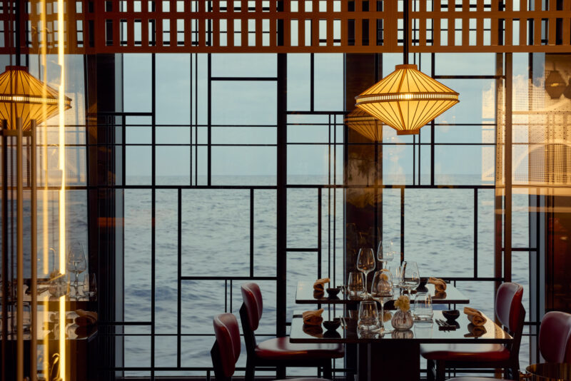 Renowned hospitality photographer Toby Mitchell for MSC Cruises’ ultra luxury travel offering Explora Journeys. An oceanview is framed by a window with unique mondrian-like mullions and soft reflections from the room's interior lighting. Beautiful lit chandeliers and dining tables are set and ready for guests in the foreground.